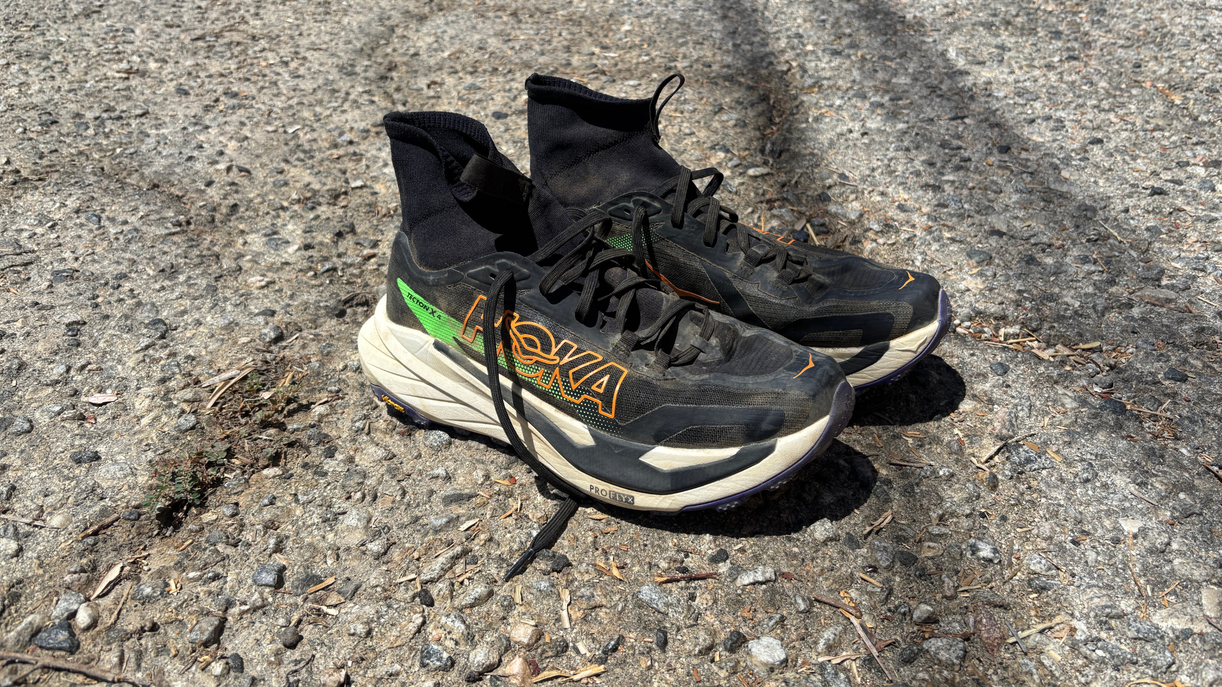 Pair of well-worn black Hoka Tecton X 4 trail running shoes with sock-like high ankle collars resting on gravel ground.